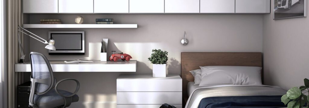 Habitación juvenil o estudio con un escritorio blanco flotante a la izquierda, una silla de oficina ergonómica, un monitor y un flexo. A la derecha hay una cama individual con cabecero de madera, una cajonera blanca intermedia y una pequeña planta.